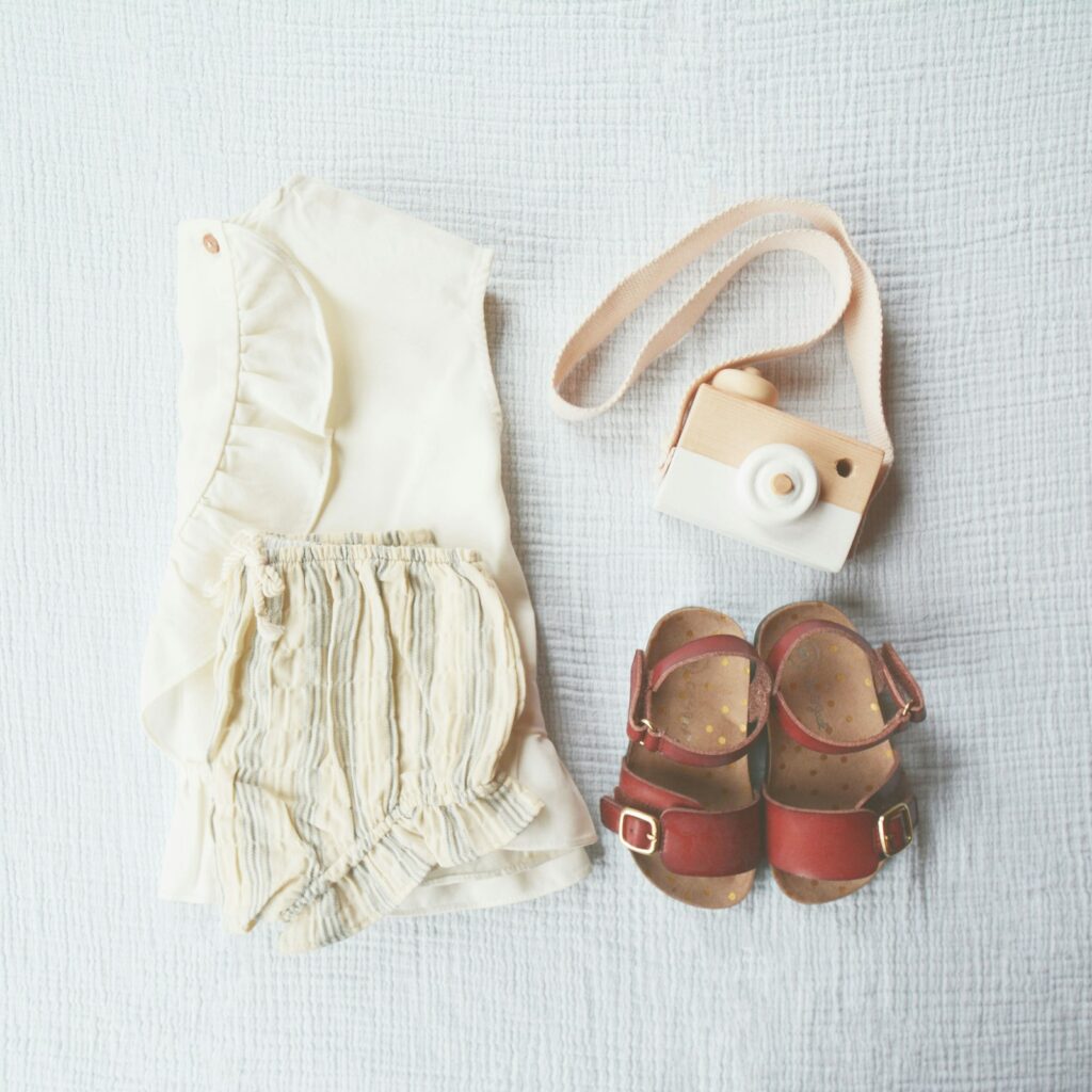 Baby clothes, sandals and a wooden toy camera arranged on a light blanket