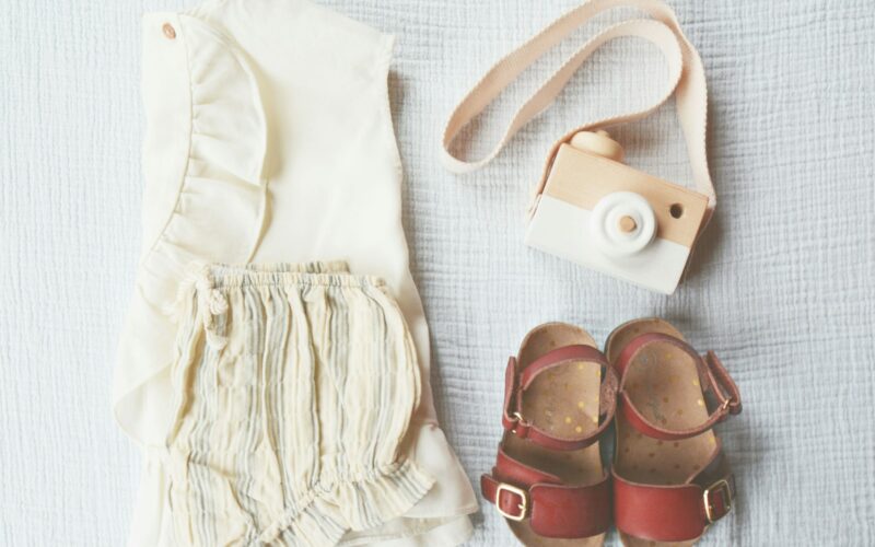 Baby clothes, sandals and a wooden toy camera arranged on a light blanket